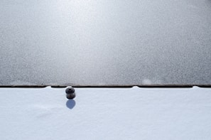 A metal bollard surrounded by fresh snow, casting a shadow on the white surface. The scene is split horizontally with a contrasting textured ground, possibly ice or pavement, above the snow.