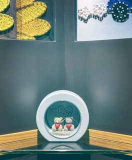 Two small teddy bears are encased together in a circular frame. One bear is cream-colored and the other is gray, each holding a red heart. The frame has the words 'Love' and 'Sweet.' Above them is a heart with multicolored dots. The background displays yellow and blue woven designs and sparkly diamond-like decorations.