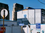 A construction site with a large, metallic building featuring the CSCEC logo and Chinese characters on it. Surrounding the building are tall apartment complexes in the background. Signs are visible, including one indicating an exit with English and Chinese text, and several informational signs in Chinese.