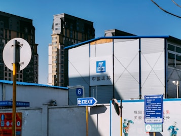 A construction site with a large, metallic building featuring the CSCEC logo and Chinese characters on it. Surrounding the building are tall apartment complexes in the background. Signs are visible, including one indicating an exit with English and Chinese text, and several informational signs in Chinese.