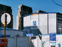 A construction site with a large, metallic building featuring the CSCEC logo and Chinese characters on it. Surrounding the building are tall apartment complexes in the background. Signs are visible, including one indicating an exit with English and Chinese text, and several informational signs in Chinese.