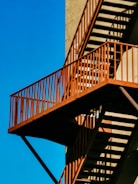 An exterior metal staircase with an industrial appearance, featuring rust-colored railings casting shadows on a beige wall. The structure stands out against a clear blue sky.