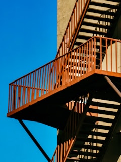 An exterior metal staircase with an industrial appearance, featuring rust-colored railings casting shadows on a beige wall. The structure stands out against a clear blue sky.