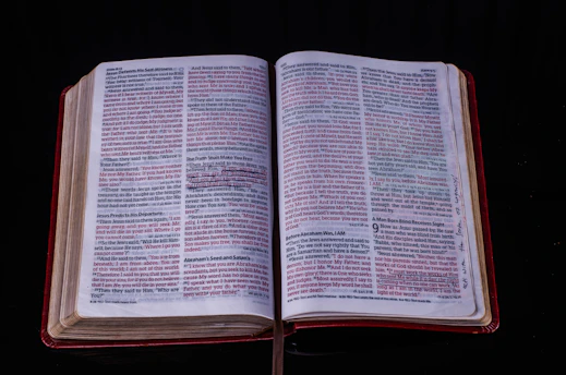 An open book displaying pages with text in two columns per page. Certain passages are highlighted in red, and there are handwritten notes in the margins. The cover is red, visible around the edges of the pages.