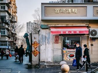 A street scene featuring a small shop with a red awning and signage in Chinese characters. Two people are walking past, one holding a blue bag and the other wearing a mask. The setting appears urban, with a tall residential building on the left, bare trees, and a clear sky in the background. Traditional red hanging decorations are seen on the shop's facade.