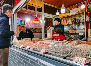 A market stall selling a variety of dried goods, including nuts and seeds, with a person wearing a red apron handling a plastic bag. Another person, likely a customer, stands in front of the stall holding a bag. The stall is decorated with colorful packaging and small red signs displaying prices.