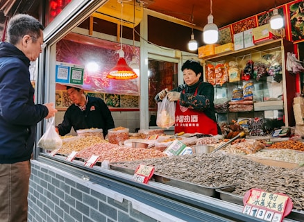 A market stall selling a variety of dried goods, including nuts and seeds, with a person wearing a red apron handling a plastic bag. Another person, likely a customer, stands in front of the stall holding a bag. The stall is decorated with colorful packaging and small red signs displaying prices.