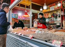A market stall selling a variety of dried goods, including nuts and seeds, with a person wearing a red apron handling a plastic bag. Another person, likely a customer, stands in front of the stall holding a bag. The stall is decorated with colorful packaging and small red signs displaying prices.