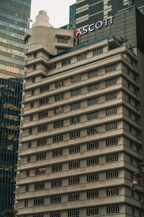 A tall, beige concrete building with many windows, each outlined by dark frames. The structure features tiered levels and a rounded corner, giving it a unique architectural design. Another modern glass building is partially visible next to it. The name 'ASCOTT' is prominently displayed near the top.