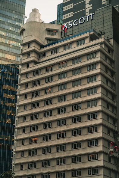 A tall, beige concrete building with many windows, each outlined by dark frames. The structure features tiered levels and a rounded corner, giving it a unique architectural design. Another modern glass building is partially visible next to it. The name 'ASCOTT' is prominently displayed near the top.
