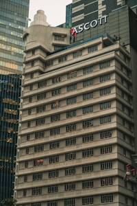 A tall, beige concrete building with many windows, each outlined by dark frames. The structure features tiered levels and a rounded corner, giving it a unique architectural design. Another modern glass building is partially visible next to it. The name 'ASCOTT' is prominently displayed near the top.