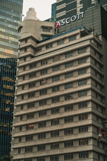 A tall, beige concrete building with many windows, each outlined by dark frames. The structure features tiered levels and a rounded corner, giving it a unique architectural design. Another modern glass building is partially visible next to it. The name 'ASCOTT' is prominently displayed near the top.
