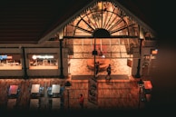 An indoor food court area with a large arched glass entrance. Two people are walking in front of the entrance. The flooring is wooden, and there are metal tables and chairs nearby. Signs for Grill Cafe and another food outlet are visible. The lighting gives a warm, inviting atmosphere.