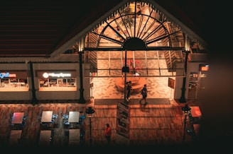 An indoor food court area with a large arched glass entrance. Two people are walking in front of the entrance. The flooring is wooden, and there are metal tables and chairs nearby. Signs for Grill Cafe and another food outlet are visible. The lighting gives a warm, inviting atmosphere.
