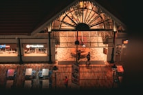 An indoor food court area with a large arched glass entrance. Two people are walking in front of the entrance. The flooring is wooden, and there are metal tables and chairs nearby. Signs for Grill Cafe and another food outlet are visible. The lighting gives a warm, inviting atmosphere.