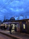 The image depicts a public transportation scene at dusk with a tram at a station. Several people are visible on the platform, some wearing masks and carrying bags. In the background, a large mosque with domes and minarets is silhouetted against a cloudy, blue-lit sky. Overhead tram wires and streetlights are also visible.