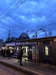 The image depicts a public transportation scene at dusk with a tram at a station. Several people are visible on the platform, some wearing masks and carrying bags. In the background, a large mosque with domes and minarets is silhouetted against a cloudy, blue-lit sky. Overhead tram wires and streetlights are also visible.