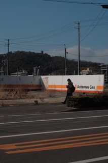 A person walking on a road beside an industrial facility with an 'Astomos Gas' sign. There are power lines above and a forested hill in the distance. The sky is clear and blue.