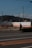 A person walking on a road beside an industrial facility with an 'Astomos Gas' sign. There are power lines above and a forested hill in the distance. The sky is clear and blue.