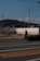 A person walking on a road beside an industrial facility with an 'Astomos Gas' sign. There are power lines above and a forested hill in the distance. The sky is clear and blue.