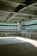 The image depicts a modern parking garage interior with concrete flooring and metal beams supporting the structure. Sunlight enters through a gap, casting soft shadows on the ground. Horizontal green stripes on a wall panel contribute to the industrial aesthetic.