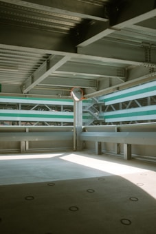 The image depicts a modern parking garage interior with concrete flooring and metal beams supporting the structure. Sunlight enters through a gap, casting soft shadows on the ground. Horizontal green stripes on a wall panel contribute to the industrial aesthetic.
