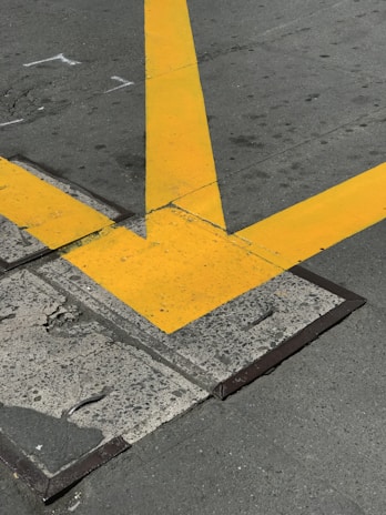A cracked concrete surface intersected by large, bold yellow lines in a V shape, indicating a traffic or parking area. The pavement shows signs of wear with visible cracks and rough textures.