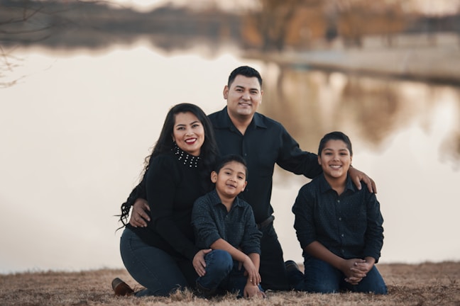 Four people are gathered together outdoors near a calm body of water, possibly a lake. They appear to be a family consisting of two adults and two children. All are dressed in dark clothing, and they are smiling, suggesting a pleasant moment. The background includes reflections of trees on the water, and the scene is set during the day.