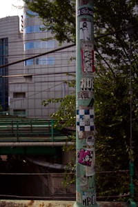 A cylindrical green pole is plastered with various stickers, including graphic designs and text. It stands in an urban environment with modern buildings in the background and a road or railway structure below. Trees with green foliage partially frame the pole.