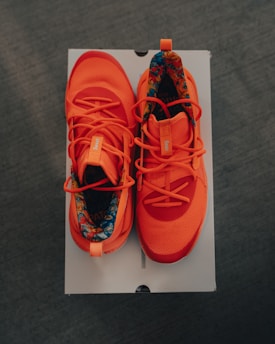 A pair of bright orange athletic shoes with patterned inner lining resting on a white rectangular box against a dark background. The shoes have intricate lace designs and tags on the tongue area.