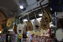 A variety of string instruments are hanging on display in a shop. The instruments include lutes with intricate patterns and some guitars. The color tones are wooden browns, and there are also bright tambourines with various colors such as red, green, and blue visible. The shop is well-lit with several spotlights and fluorescent lights illuminating the instruments.