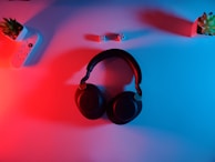 A pair of black over-ear headphones placed on a surface illuminated with red and blue lighting. Surrounding the headphones, there are two small potted plants, one on each side, and a remote control with buttons on the left side. Two white dice with black dots are placed slightly above the headphones.