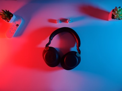 A pair of black over-ear headphones placed on a surface illuminated with red and blue lighting. Surrounding the headphones, there are two small potted plants, one on each side, and a remote control with buttons on the left side. Two white dice with black dots are placed slightly above the headphones.