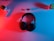 A pair of black over-ear headphones placed on a surface illuminated with red and blue lighting. Surrounding the headphones, there are two small potted plants, one on each side, and a remote control with buttons on the left side. Two white dice with black dots are placed slightly above the headphones.