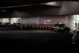 Several taxis are lined up under a dimly lit urban structure. The area is characterized by a series of illuminated bollards, part of an organized taxi queue outside a building with reflective surfaces. People can be seen in the background near the entrance of a brightly lit area.