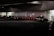 Several taxis are lined up under a dimly lit urban structure. The area is characterized by a series of illuminated bollards, part of an organized taxi queue outside a building with reflective surfaces. People can be seen in the background near the entrance of a brightly lit area.
