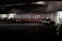 Several taxis are lined up under a dimly lit urban structure. The area is characterized by a series of illuminated bollards, part of an organized taxi queue outside a building with reflective surfaces. People can be seen in the background near the entrance of a brightly lit area.