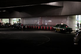 Several taxis are lined up under a dimly lit urban structure. The area is characterized by a series of illuminated bollards, part of an organized taxi queue outside a building with reflective surfaces. People can be seen in the background near the entrance of a brightly lit area.