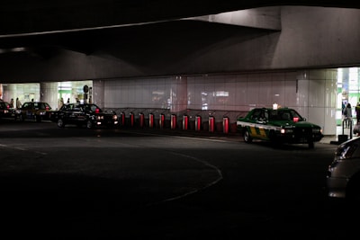 Several taxis are lined up under a dimly lit urban structure. The area is characterized by a series of illuminated bollards, part of an organized taxi queue outside a building with reflective surfaces. People can be seen in the background near the entrance of a brightly lit area.
