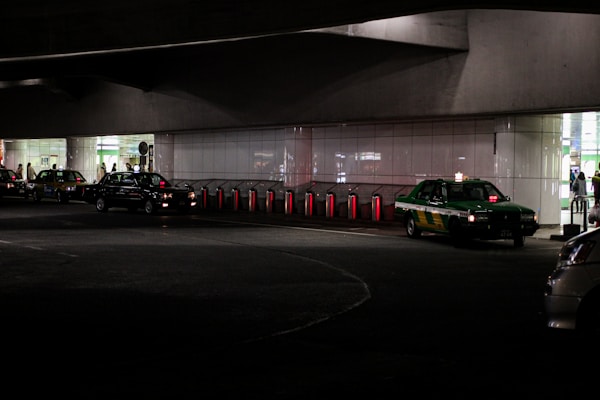 Several taxis are lined up under a dimly lit urban structure. The area is characterized by a series of illuminated bollards, part of an organized taxi queue outside a building with reflective surfaces. People can be seen in the background near the entrance of a brightly lit area.