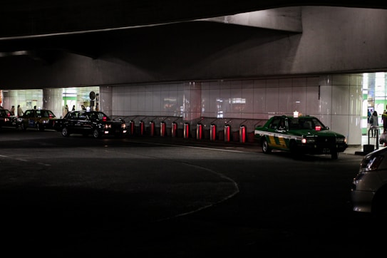 Several taxis are lined up under a dimly lit urban structure. The area is characterized by a series of illuminated bollards, part of an organized taxi queue outside a building with reflective surfaces. People can be seen in the background near the entrance of a brightly lit area.
