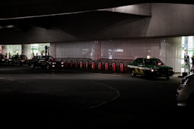 Several taxis are lined up under a dimly lit urban structure. The area is characterized by a series of illuminated bollards, part of an organized taxi queue outside a building with reflective surfaces. People can be seen in the background near the entrance of a brightly lit area.
