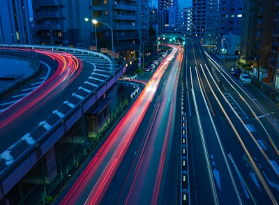 A nighttime cityscape featuring elevated roads with light trails from traffic, indicating fast movement. The roads curve and intersect among tall buildings illuminated with city lights.