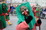 A street parade scene features several participants wearing colorful costumes and traditional masks. The masks have exaggerated facial features such as large eyes and noses. The central figure is wearing a green and red outfit, and is holding a stick. A crowd observes the parade from behind metal barriers in the background.