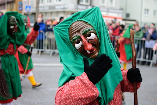 A street parade scene features several participants wearing colorful costumes and traditional masks. The masks have exaggerated facial features such as large eyes and noses. The central figure is wearing a green and red outfit, and is holding a stick. A crowd observes the parade from behind metal barriers in the background.