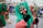A street parade scene features several participants wearing colorful costumes and traditional masks. The masks have exaggerated facial features such as large eyes and noses. The central figure is wearing a green and red outfit, and is holding a stick. A crowd observes the parade from behind metal barriers in the background.
