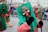 A street parade scene features several participants wearing colorful costumes and traditional masks. The masks have exaggerated facial features such as large eyes and noses. The central figure is wearing a green and red outfit, and is holding a stick. A crowd observes the parade from behind metal barriers in the background.