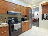 A modern kitchen with wooden cabinets and a stainless steel oven. A kettle sits on the stovetop, and a microwave is on the countertop. The kitchen has a light color scheme with beige tiles and a partial view into a living room with a black and orange color palette.