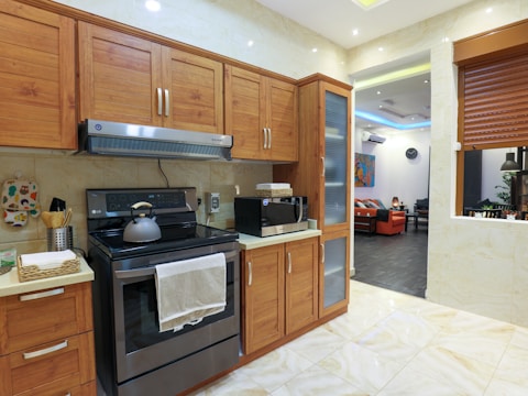 A modern kitchen with wooden cabinets and a stainless steel oven. A kettle sits on the stovetop, and a microwave is on the countertop. The kitchen has a light color scheme with beige tiles and a partial view into a living room with a black and orange color palette.