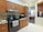 A modern kitchen with wooden cabinets and a stainless steel oven. A kettle sits on the stovetop, and a microwave is on the countertop. The kitchen has a light color scheme with beige tiles and a partial view into a living room with a black and orange color palette.